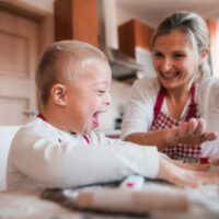 A laughing down syndrome child with his mother indoors baking.