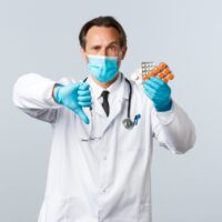Covid-19, preventing virus, healthcare workers and vaccination concept. Displeased serious doctor in medical mask and gloves, show thumb-down and medication, bad quality pills, wrong prescription Covid-19, preventing virus, healthcare workers and vaccination concept. Displeased serious doctor in medical mask and gloves, show thumb-down and medication, bad quality pills, wrong prescription
