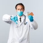 Covid-19, preventing virus, healthcare workers and vaccination concept. Displeased serious doctor in medical mask and gloves, show thumb-down and medication, bad quality pills, wrong prescription Covid-19, preventing virus, healthcare workers and vaccination concept. Displeased serious doctor in medical mask and gloves, show thumb-down and medication, bad quality pills, wrong prescription