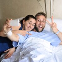 young couple taking selfie photo at hospital room with man in bed