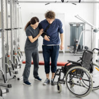 Rehabilitation specialist helps a guy stand out of a wheelchair at rehabilitation center. Concept of physical therapy and support for people with disabilities