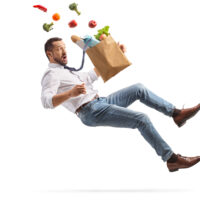 A man in a white shirt and jeans, levitating, with a surprised expression. A paper grocery bag spills vegetables, fruit, and bread around him.