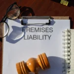 A clipboard with a paper titled "Premises Liability" sits on a wooden desk with glasses, a judge's gavel, a notebook, and a pen, conveying a legal theme.